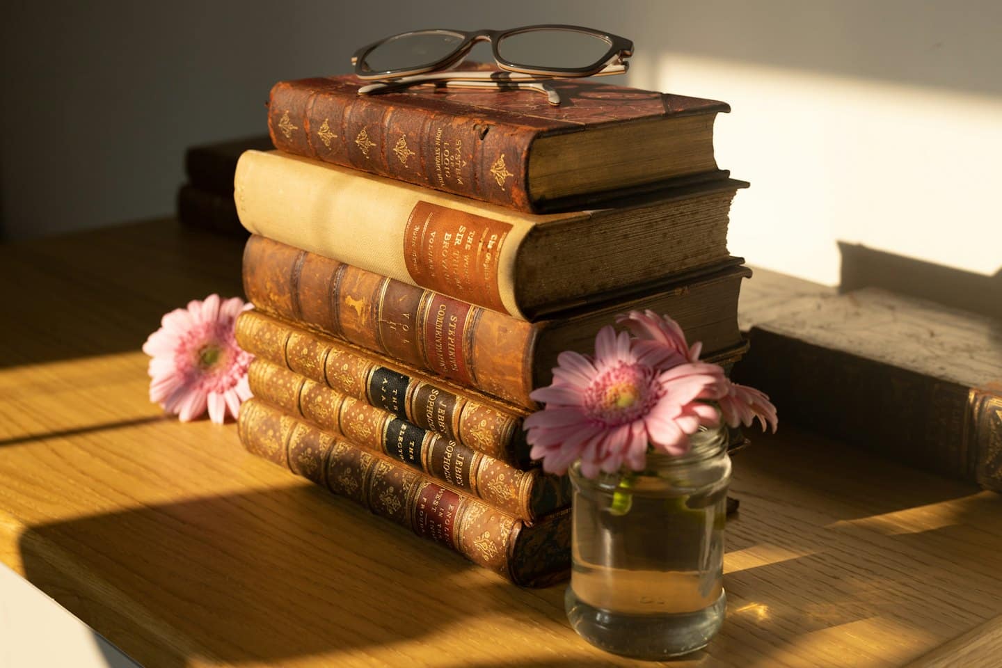 Libri antichi con occhiali da lettura e margherite rosa su legno.