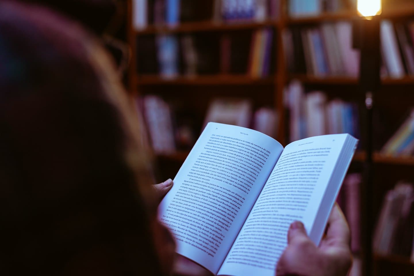 Persona che legge un libro aperto con librerie sfocate alle spalle.